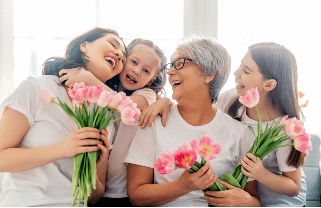 Two women and two girls hugging with flowers in hand, smiling and sharing a joyful moment across generations.